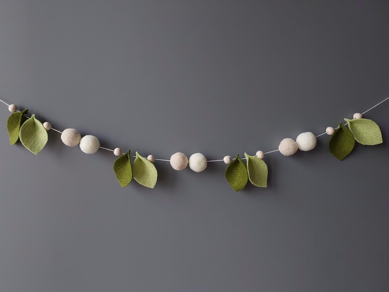 May include: A decorative garland featuring felt leaves and round, light-colored beads. The leaves are a soft green, and the beads are a mix of white and beige. The garland is strung on a thin white cord, perfect for home decor.