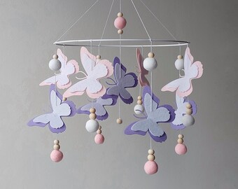 Könnte beinhalten: Ein Baby-Mobile mit mehrlagigen Papier-Schmetterlingen in den Farbtönen Lavendel, Rosa und Weiß. Die Schmetterlinge sind an einem weißen Ring mit Holzperlen und rosa und weißen Filzkugeln aufgehängt. Ein dekorativer Artikel für das Kinderzimmer.