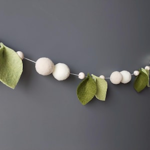 May include: A decorative garland featuring felt leaves in shades of green and white felt balls. The garland is strung on a thin white cord, creating a whimsical and natural aesthetic. Perfect for adding a touch of nature to any space.
