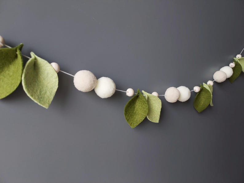 May include: A decorative garland featuring felt leaves in shades of green and white felt balls. The garland is strung on a thin white cord, creating a whimsical and natural aesthetic. Perfect for adding a touch of nature to any space.