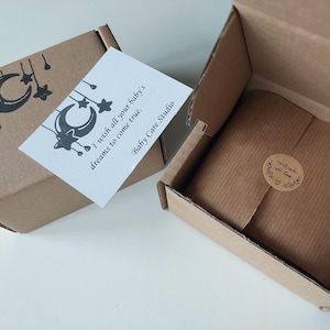 May include: Two open cardboard boxes with a baby-themed gift. One box has a card that reads "I wish all your baby's dreams to come true. Baby Care Studio." The other box contains a wrapped gift with a "handmade with love" sticker.