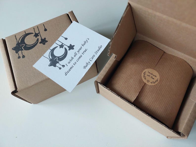May include: Two open cardboard boxes with a baby-themed gift. One box has a card that reads "I wish all your baby's dreams to come true. Baby Care Studio." The other box contains a wrapped gift with a "handmade with love" sticker.