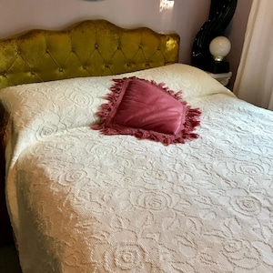 May include: A bed features a tufted mustard yellow velvet headboard and a white bedspread with a raised floral rose pattern. A square, dusty rose-colored throw pillow with a fringed edge sits in the center of the bedspread.