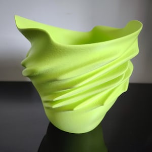 Könnte beinhalten: Eine limettengrüne Keramikvase mit einem strukturierten, gewellten Design. Die Vase hat eine abstrakte, organische Form mit einem ausgestellten Oberteil und einem glatten Sockel. Es ist ein dekorativer Gegenstand.