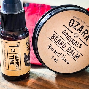 May include: Two beard care products, a 1 oz. bottle of Harvest Time beard oil and a 2 oz. tin of Harvest Time beard balm, both from Ozark Originals. The products are on a wooden surface with a red plaid fabric in the background.