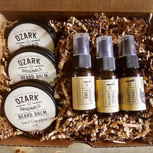 Może przedstawiać: Pudełko prezentowe wypełnione produktami do pielęgnacji brody. Trzy małe buteleczki olejku do brody z etykietami z napisem "Harvest Time", "Forest Campfire" i "Tobacco & Coffee". Trzy małe słoiczki balsamu do brody z etykietami z napisem "Ozark Originals Beard Balm" i "Ozark Originals Beard Balm Forest Campfire 2 oz."