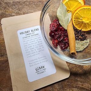 Könnte beinhalten: Eine Glasschüssel mit Potpourri-Zutaten, darunter getrocknete Cranberries, Orangenscheiben, Zimtstange, Nelken und Rosmarin. Daneben ein Kraftpapierbeutel mit der Aufschrift "HOLIDAY BLEND" und "OZARK ORIGINALS".