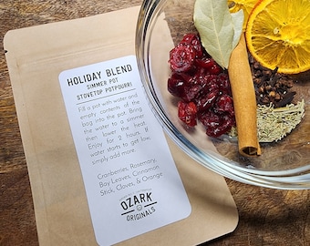 May include: A glass bowl containing potpourri ingredients: dried cranberries, orange slices, a cinnamon stick, cloves, and rosemary. A kraft paper bag with the text "HOLIDAY BLEND" and "OZARK ORIGINALS" is next to the bowl.