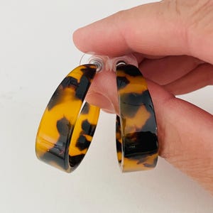 May include: A pair of tortoise shell hoop earrings. The earrings feature a classic hoop design with a rounded shape. The earrings have a warm, brown and black pattern.