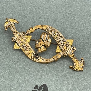 May include: A vintage gold-toned brooch featuring a central profile of a person with a head wrap, framed by two crossed swords. The swords and frame are adorned with intricate floral and geometric patterns.