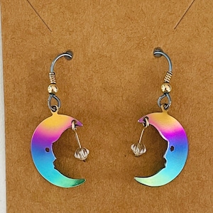 May include: A pair of dangle earrings featuring crescent moon charms with a gradient rainbow finish. Each moon charm is suspended from a silver-toned hook and accented with a small, clear, faceted bead hanging from the inner curve of the moon.