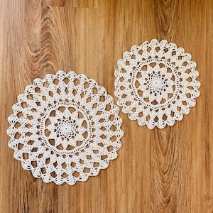 May include: Two off-white, crocheted doilies with intricate, circular patterns. The doilies are placed on a wooden surface, showcasing their delicate, openwork design. The handmade items are ideal for home decor.