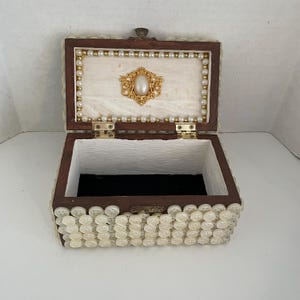 May include: An open, vintage jewelry box with a dark brown wooden frame. The box is decorated with small, white buttons and a pearl and gold-toned trim. The interior features a black velvet base and a gold-toned embellishment.