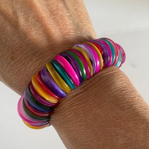 May include: A colorful bracelet made of stacked, round, iridescent shell pieces. The bracelet is a mix of pink, purple, green, yellow, and orange.