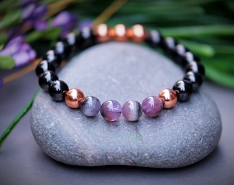 May include: A beaded bracelet with black, copper, and purple beads. The bracelet is resting on a gray stone.