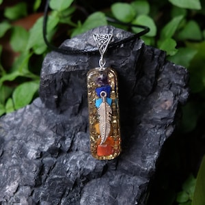 May include: A silver pendant with a clear resin rectangle containing seven chakra stones and a silver feather charm. The pendant is on a black cord and is resting on a dark gray rock.