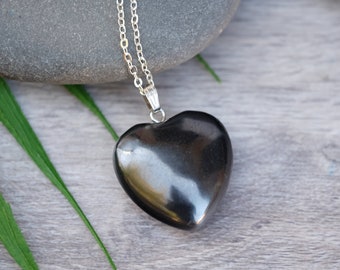 May include: A heart-shaped, polished black pendant necklace with a silver chain. The pendant is smooth and reflective, set against a backdrop of gray stones and wooden planks, with green leaves.