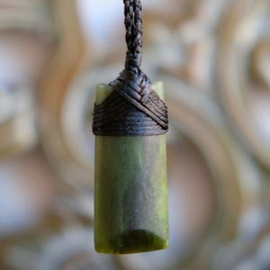 Könnte beinhalten: Ein grüner Jade-Anhänger mit zylindrischer Form, der an einer dunkelbraunen geflochtenen Kordel hängt. Die Kordel ist um die Oberseite des Anhängers gewickelt. Der Hintergrund ist verschwommen, mit einem hellbraunen, verzierten Muster.