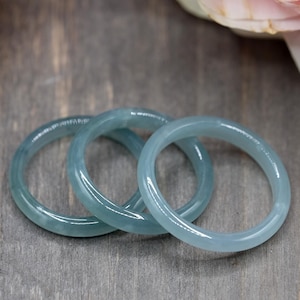 Könnte beinhalten: Drei hellblaue Jade-Armreifen, die auf einer Holzoberfläche gestapelt sind. Die Armreifen sind glatt und poliert.
