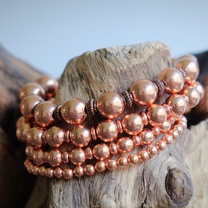 May include: Four copper beaded bracelets of varying sizes are displayed on a piece of weathered wood. The bracelets have a warm, metallic sheen, with the beads ranging from small to large. The wood has a natural, textured appearance, with a light brown color.