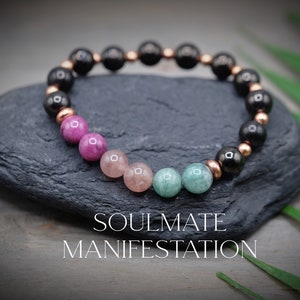 Puede incluir: Una pulsera de cuentas con piedras preciosas negras, rosas, melocotón y verdes, intercaladas con cuentas de color cobre. La pulsera se muestra sobre una piedra gris oscura con el texto "SOULMATE MANIFESTATION" en blanco.