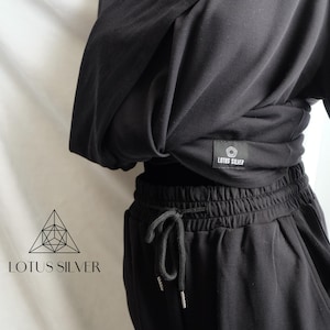 Könnte beinhalten: Schwarzes zweiteiliges Sport-Set. Das Oberteil ist ein Wickelstil mit langen Ärmeln und einem kleinen Etikett mit der Aufschrift „LOTUS SILVER“. Die Hose ist eine Jogginghose mit Kordelzug. Das Set ist aus einem weichen, dunklen Stoff gefertigt.