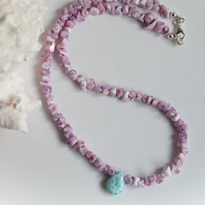 May include: A delicate necklace featuring small, light purple shell beads. The necklace has a silver clasp and a single, light blue teardrop-shaped pendant. A large, white seashell is in the background.