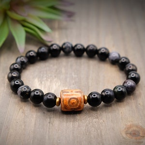 May include: Black beaded bracelet with a brown and orange carved bead in the center.