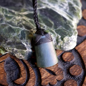 Könnte beinhalten: Ein dunkelgrüner Jade-Anhänger mit einer schwarzen geflochtenen Kordel. Der Anhänger hat eine glatte, abgerundete Form und wird vor einem strukturierten grünen Stein und einer geschnitzten Holzoberfläche präsentiert.