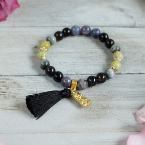 May include: A beaded bracelet featuring a mix of black, gray, yellow, and blue-purple beads. A black tassel and a gold-colored charm hang from the bracelet. The bracelet is displayed on a white wooden surface with pink rose petals in the background.