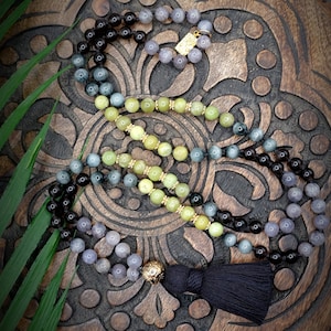 May include: A multi-strand mala necklace with black, gray, and green beads. The necklace has a tassel at the end and is laid out on a wooden surface.