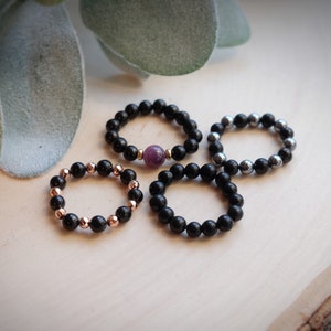 May include: Four beaded rings, three black and one with a purple gemstone. The rings are on a wooden surface.