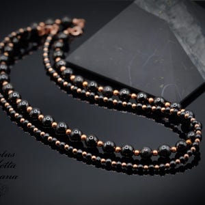 May include: A layered necklace featuring black beads and smaller copper-colored beads. The necklace is displayed on a reflective black surface, with a dark background. The necklace has a copper-colored clasp.