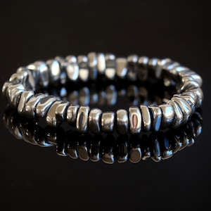 May include: A silver-colored bracelet composed of numerous small, irregularly shaped beads. The beads are strung together to form a flexible band. The bracelet is displayed on a reflective black surface, enhancing its metallic sheen.