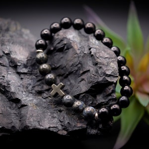 May include: Black beaded bracelet with a silver cross charm, resting on a dark gray rock. The bracelet is made of round beads and has a simple design.