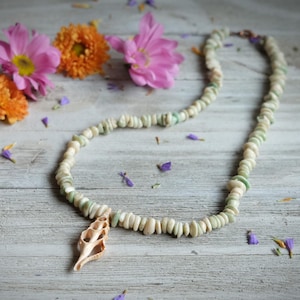 May include: A handmade necklace featuring a strand of small, off-white and green beads. A natural seashell pendant hangs from the center. The necklace is displayed on a light wood surface, with pink and orange flowers in the background.
