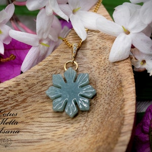 May include: A jade green snowflake pendant necklace with a gold chain and small clear crystals. The necklace is displayed on a wooden surface with white and pink flowers.