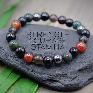 May include: A beaded bracelet with black, red, green, and grey stones. The bracelet has the words "STRENGTH COURAGE STAMINA" printed on it.