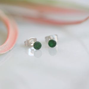 May include: A pair of stud earrings with small, round, dark green gems set in silver posts. The earrings are displayed on a white surface with a blurred pink and white background.