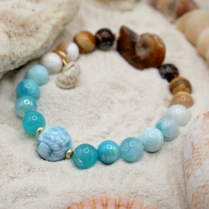May include: A blue and white gemstone bracelet with a carved turtle charm. The bracelet is made with round beads and is displayed on a white sandy surface with seashells.