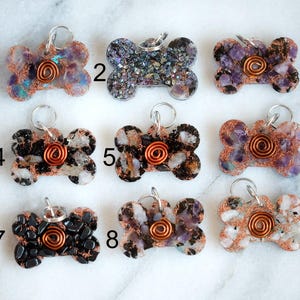 May include: Nine dog bone-shaped pet tags with a copper wire spiral in the centre. The tags are made of resin and embedded with various stones and crystals in shades of purple, black, and white. Each tag has a silver-coloured ring for attaching to a collar.