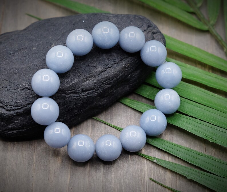 May include: A light blue gemstone bracelet with round beads. The bracelet is on a dark grey stone and green leaves.