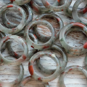 May include: A collection of smooth, circular rings crafted from translucent pale green stone featuring distinct, irregular inclusions of opaque red and dark grey mineral deposits throughout the polished surface of each individual band.