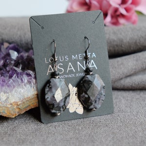 May include: A pair of dangle earrings with faceted, oval-shaped, gray and black stones. The earrings are suspended from black metal hooks and feature small black beads. The earrings are displayed on a gray card with the text "LOTUS METTA ASANA HANDMADE JEWELRY".
