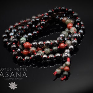 May include: A black and red beaded mala necklace with a lotus flower charm. The beads are made of hematite, red jasper, and green aventurine. The necklace is handmade and is called "LOTUS METTA ASANA".