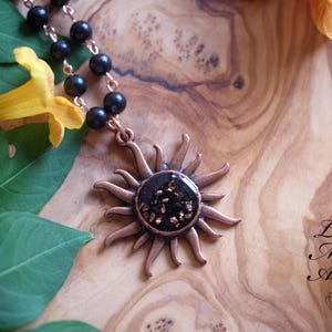 May include: A necklace featuring a copper-toned sun pendant with a dark, textured center and a chain of round black beads. The text "Lotus Metta Asana" and a lotus flower icon are printed in the lower right corner of the image.