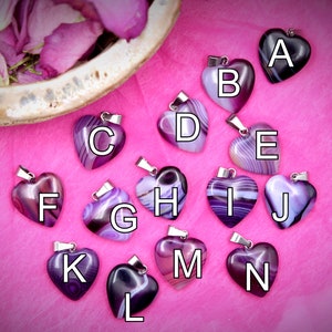 May include: Thirteen purple agate heart pendants with silver bail, labeled A through N. The pendants are arranged in a semi-circle on a pink surface.