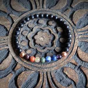 May include: A black beaded bracelet with seven colorful gemstone beads in the center. The bracelet is on a wooden surface with a carved design.