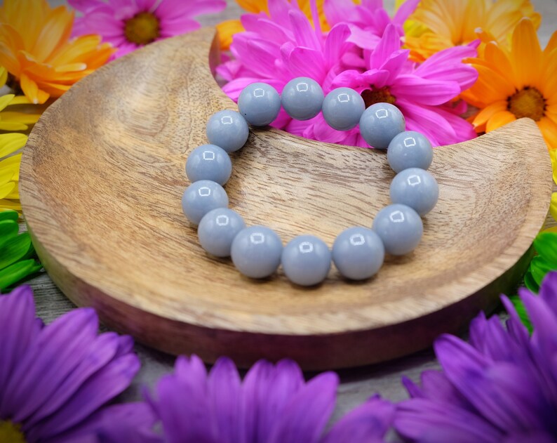 May include: A grey gemstone bracelet with round beads on a wooden crescent moon-shaped dish. The dish is surrounded by colourful flowers.