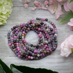 May include: A close-up of a multi-colored beaded bracelet made with pink, purple, and gray stones. The bracelet is made of many strands of beads, creating a layered effect.